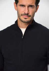Maglione zip-up nero con colletto alto, realizzato in un morbido materiale a coste. Presenta una cerniera argentata e un design aderente.