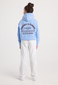 Donna con capelli ricci raccolti in un bun, che indossa un hoodie azzurro chiaro con un testo motivazionale e pantaloni della tuta bianchi, in piedi davanti a uno sfondo semplice.