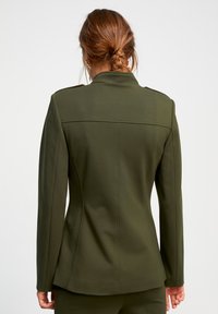 Blazer vert olive avec une coupe ajustée, un design d'épaules structurées, une fente au dos et une texture de tissu lisse. Pas de quincaillerie visible.