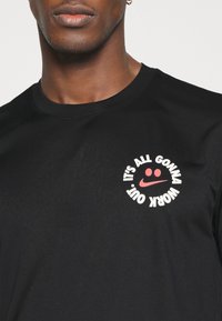 Camiseta de algodón negra con un prominente gráfico circular que presenta la frase "TODO VA A SALIR BIEN" en blanco y rojo, incluyendo una carita sonriente.