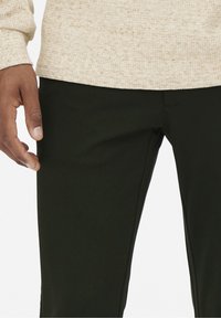 Dark green pants made from textured fabric, featuring a slim fit and a flat front. Hand positioned on the waistband.