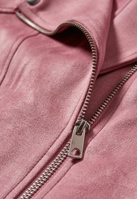 Różowa zamszowa kurtka z widocznym zamkiem błyskawicznym i srebrnymi akcentami. Miękka tekstura i gładkie wykończenie podkreślają zorganizowane elementy projektu.