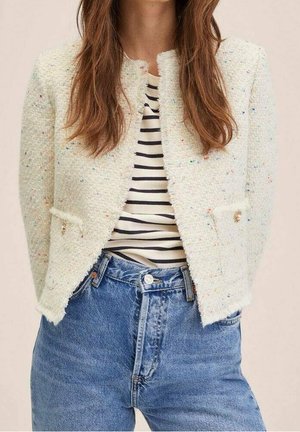 Femme portant une veste courte crème texturée sur une chemise rayée noir et blanc avec un jean bleu taille haute.