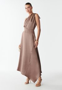 Abito maxi in satin marrone con orlo asimmetrico, design a spalla unica e dettagli annodati. Il modello tiene una pochette rotonda in metallo e indossa dei sandali con cinturini.