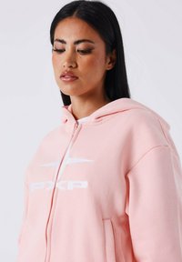 Roze hoodie met een ritssluiting, voorzien van een wit logo en een zachte, gestructureerde stof. Inclusief zakken aan de voorkant en een capuchon.