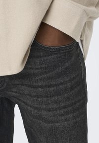 Los jeans de mezclilla gris oscuro presentan un ajuste clásico con una textura suave y ligeramente desgastada. Una capa de sudadera beige es visible en la parte superior.