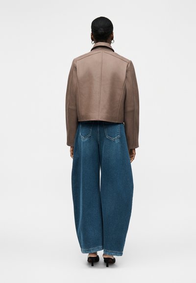 Γυναίκα με μαλλιά σε κότσο, που φορά ένα καφέ κοντό μπουφάν, φαρδιά μπλε τζιν και μαύρα παπούτσια με τακούνι, στέκεται με την πλάτη προς τα έξω σε λευκό φόντο.