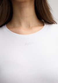 Weißes Baumwoll-T-Shirt mit Rundhalsausschnitt, das ein dekoratives gepunktetes Logo in der oberen linken Ecke aufweist, körpernahe Passform und glatte Textur.