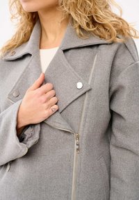 Szary płaszcz z wełny z domieszką, z klapami, srebrnymi akcentami i skośnym zamkiem błyskawicznym z przodu. Widoczna miękka tekstura.