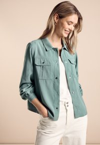 Zelená bunda na knoflíky s dvěma náprsními kapsami, měkký materiál a ohrnuté rukávy. Nositá přes bílý top a světlé kalhoty.
