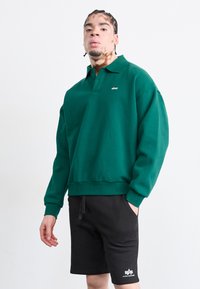 Sweatshirt polo en coton vert à manches longues, avec des poignets côtelés et un petit logo. Associé à un short noir avec des poches latérales et un détail de logo.