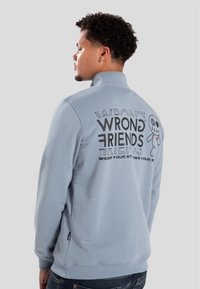 Ljusblå sweatshirt med hög krage, med svart text och en tecknad karaktärs grafik på baksidan. Mjuk kvalitetsmaterial, avslappnad design.