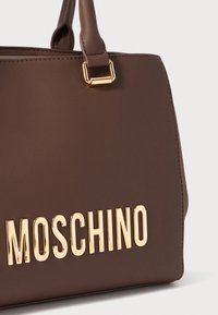 Prune nahkakott, millel on struktureeritud kuju, kuldset värvi riistvara ja esiküljel reljeefne "MOSCHINO" logo, rõhutades disainielemente.