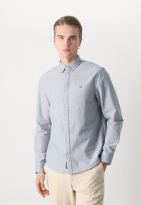 Μπλε πουκάμισο με μακριά μανίκια και κουμπιά, με γιακά, που διαθέτει ένα μικρό λογότυπο στο στήθος, φτιαγμένο από υλικό με υφή. Συνδυασμένο με ανοιχτόχρωμο παντελόνι.