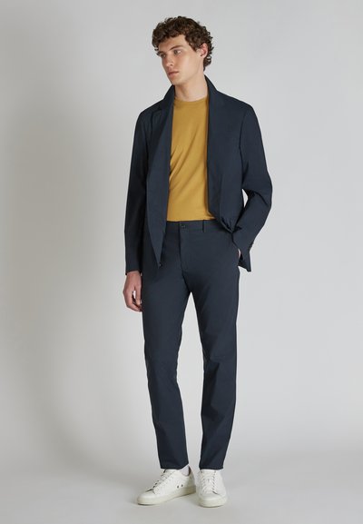 Giacca blu scuro sopra una t-shirt senape, abbinata a pantaloni navy slim-fit e sneakers bianche. Tessuto liscio, design moderno.