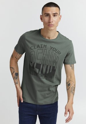 Grøn bomulds T-shirt med rund hals. Har fed sort tekst: "CLAIM YOUR DENIM." Båret med blå jeans, der viser arm tatoveringer.