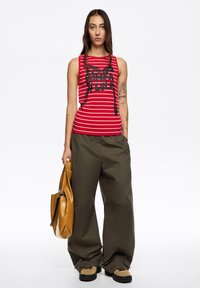Czerwono-biała pasiasta koszulka na ramiączkach z czarnym nadrukiem, zestawiona z oliwkowozielonymi, luźnymi spodniami. Trzyma brązową skórzaną torbę typu tote. Czarne buty.