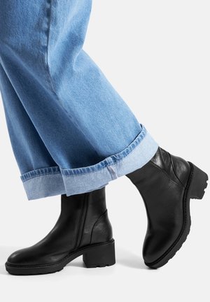 Czarne skórzane botki za kostkę z okrągłym noskiem i masywnym obcasem, zestawione z podwiniętymi jasnoniebieskimi dżinsami. Teksturowana powierzchnia i widoczny detal zamka błyskawicznego.