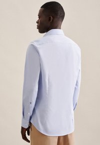 Lyseblå langermet skjorte laget av mykt stoff. Har spiss krage, knapper ved ermene, og buet nederkant med subtile bakpermer.