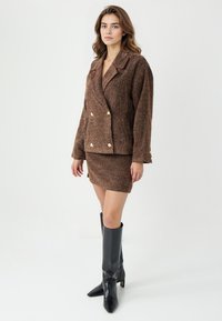 Bruine tweed dubbelzijdige blazer met gouden knopen, gecombineerd met een aansluitende bruine tweed rok. Model draagt kniehoge zwarte hakken.