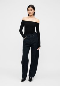 Fekete vállat szabadon hagyó hosszú ujjú felső magas derekú, sötétkék, bőszárú nadrággal párosítva. A modell zsebében tartja a kezét, fekete cipőt visel.