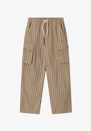 Kind mit lockigem Haar, das einen gelben Pullover und gestreifte Cargohosen trägt, balanciert mit ausgestreckten Armen vor einem beigen Hintergrund.