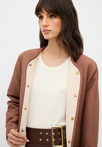 Brun quiltad jacka med gyllene tryckknappar över en krämfärgad ribbad topp och bruna högmidjade byxor med bälte. Släta texturer och lagerdesign.