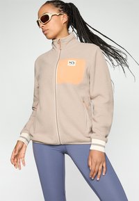 Beige fleecejakke med lynlås foran, orange brystlomme, hvid piping og stribede ærmer. Båret med tætsiddende blå leggings.