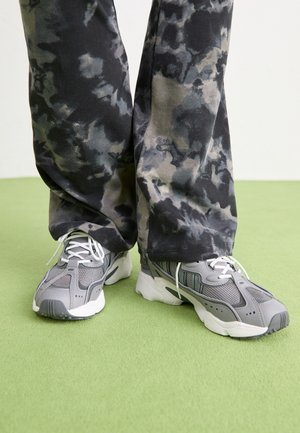 Stopy w szaro-białych sportowych butach i luźnych, czarno-szarych spodniach w stylu tie-dye stoją na zielonym dywanie.