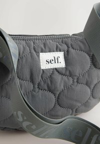 Pikowana szara torba z tkaniny z przeszywanym wzorem, biała etykieta z napisem „self.” oraz pasujący szary pasek z powtarzającym się nadrukiem „self.”.