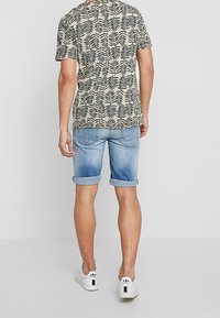 Kortärmad beige skjorta med svart lövmönster, kombinerad med ljusblå denimshorts med uppvikt fåll och vita sneakers.