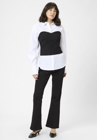 Sort korset top over en hvid skjorte med knapper, parret med sorte flagrende bukser og spidse sorte sko. Glat stof med struktureret design.