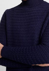 Granatowy sweter dzianinowy o teksturowanym wzorze, z prążkowanym kołnierzem i mankietami. Materiał wydaje się miękki i przytulny.