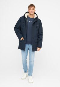 Parka de color azul marino, aislada, con capucha forrada de piel; cuenta con bolsillos frontales y cierre de cremallera. Llevado con jeans de color azul claro y zapatillas blancas.