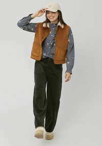 Gilet en suède marron avec un col crème, associé à une chemise bleue à motifs et un pantalon en velours côtelé vert foncé. Des chaussures beige complètent le look.
