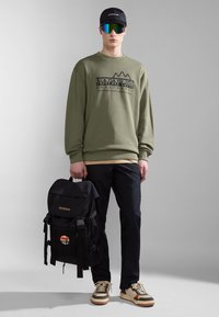 Felpa verde con logo, cappellino nero, occhiali da sole riflettenti, zaino nero, pantaloni scuri e scarpe beige con dettagli marroni.