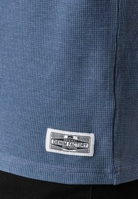 Blått strukturerat tyg med ribbat mönster, med en vit etikett som läser "ORIGINAL DENIM FACTORY" nära botten.