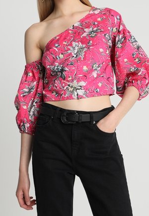 Vrouw draagt een roze bloemige asymmetrische crop top met pofmouwen en zwarte hooggetailleerde broek met riem, hand in de zak.