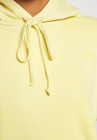 Žlutá mikina s kapucí a stahovací šňůrkou, ribovaná textura a vyšitým logem na hrudi. Mělo by se zdát, že látka je měkká a lehká.