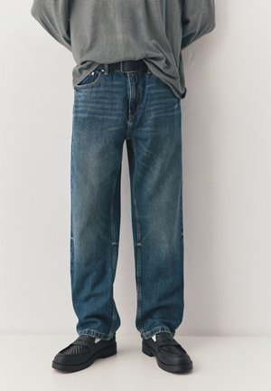 Person trägt lockere blaue Jeans, schwarzen Gürtel, schwarze Loafers mit weißen Socken und ein graues, gestreiftes Langarmhemd, vorne eingesteckt.