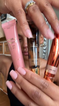 Tre tubetti cosmetici tenuti in mano: un tubetto rosa con etichetta "Hydra Bright", un tubetto marrone scuro "Cream Contour & Bronze" e un tubetto arancione metallizzato "Ablaze".