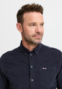 Marineblauwe corduroy button-up shirt met kraag, voorzak met een kleine driekleurige patch, en twee knopen op de kraag.