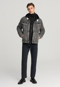 Giacca di denim con colletto morbido a contrasto, indossata sopra un hoodie nero con zip. Jeans e scarpe neri. In generale, un outfit monochrome a strati.