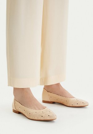 Stopy w beżowych, niskich balerinach z kwiatowymi wycięciami, w połączeniu z kremowymi spodniami o szerokich nogawkach na jednolitym tle.