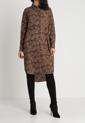 Femme portant une robe imprimée léopard à manches longues et boutonnée, associée à des bottes noires à talons hauts au-dessus du genou, debout devant un fond uni.