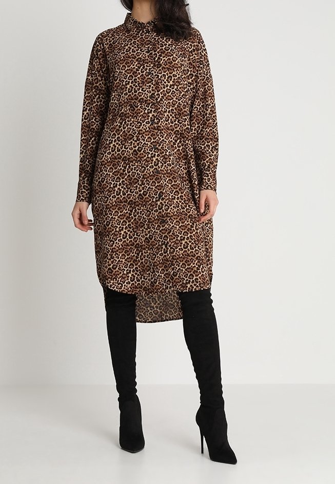 Femme portant une robe imprimée léopard à manches longues et boutonnée, associée à des bottes noires à talons hauts au-dessus du genou, debout devant un fond uni.