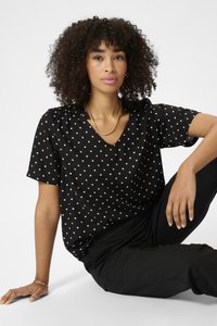 Schwarze Bluse mit weißen Punkten, V-Ausschnitt, kurzen Ärmeln und gerafften Schulterdetails. Getragen mit schwarzen Hosen.