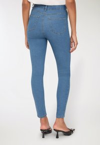 Kobieta w obcisłych niebieskich dżinsach i czarnych klapkach na obcasie, stojąca tyłem do kamery na jednolitym białym tle.