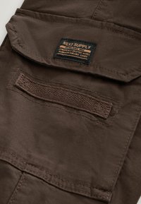 Brown fabric pocket with flap and woven label reading "NEXT SUPPLY Established 1982 Authentically Crafted Wares Trademark Durable Goods."