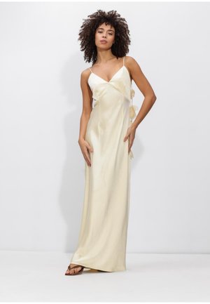 Femme aux cheveux bouclés portant une longue robe nuisette en satin crème à fines bretelles et des sandales brunes, debout contre un mur blanc.